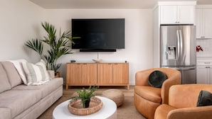 Una sala moderna con sofá, sillón y mesa de centro, un televisor de pantalla plana montado en la pared y una zona de cocina con refrigerador.