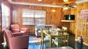Een gezellige kamer met houten wanden, een eettafel gedekt voor vier personen, twee roze stoelen en een grijze bank.