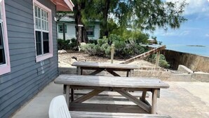 Een terras aan het strand met een houten picknicktafel en twee witte lounge fauteuils.