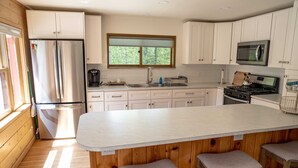 A modern kitchen with white cabinets, a stainless steel refrigerator, a microwave, and a gas stove. There is a large window providing natural light and a wooden island with bar stools.