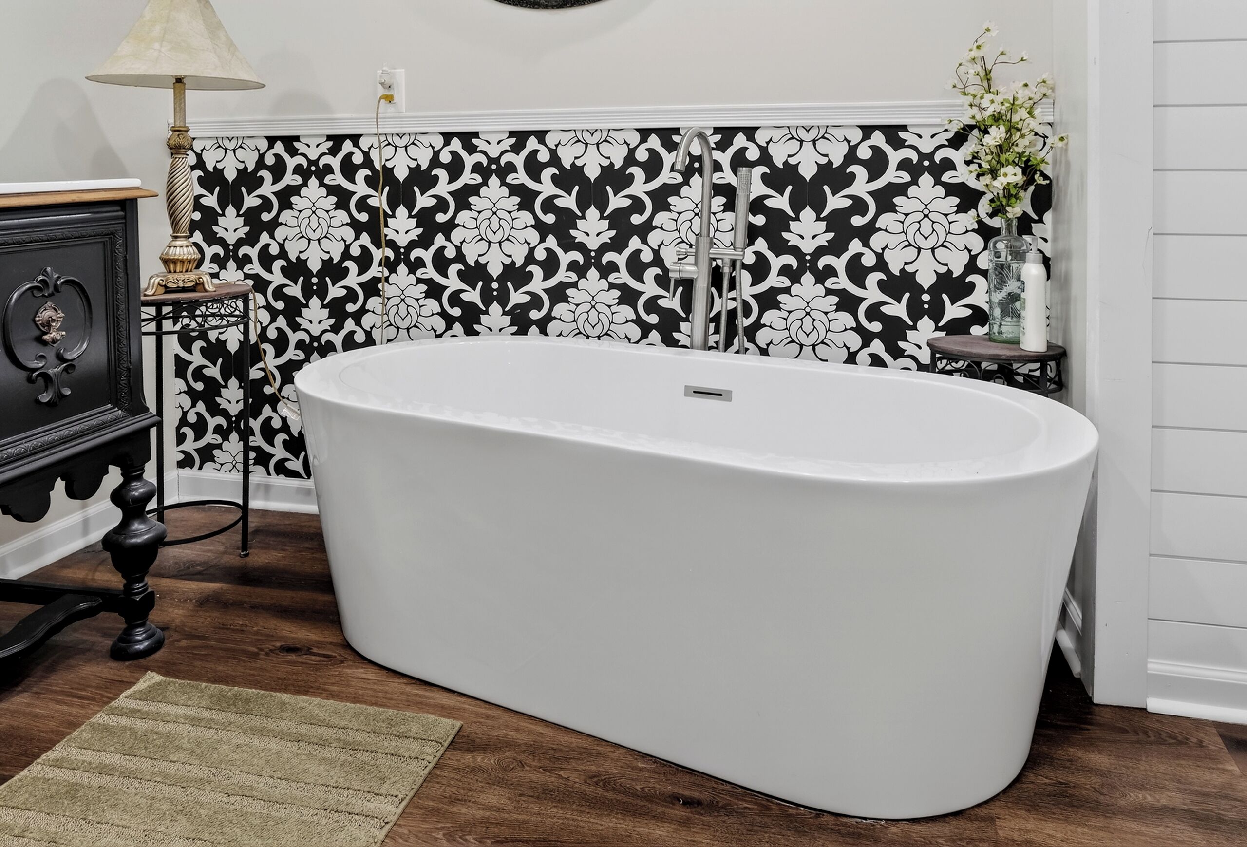 A bathroom with a freestanding bathtub, a black and white patterned wall, a wooden floor, a small table with a lamp, and a vase with flowers.