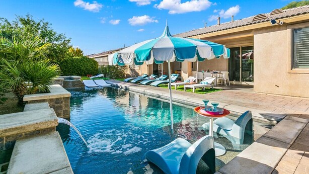 Ein Schwimmbecken mit Liegestühlen und Sonnenschirmen, einer kleinen Wasserfallanlage und einer Terrassenbereiches mit Gartenmöbeln.