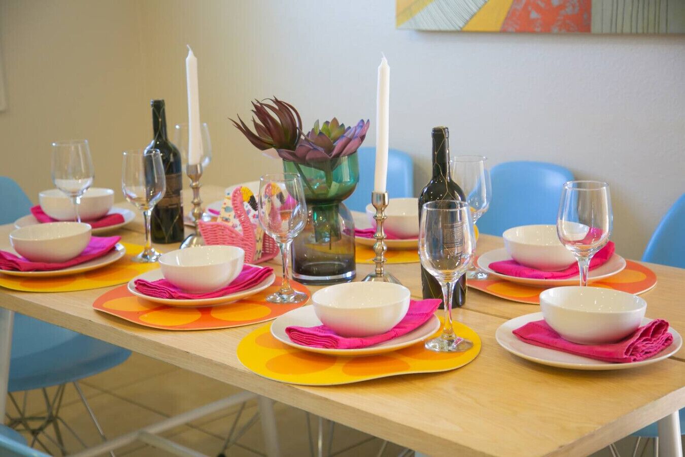 Mesa de comedor dispuesta con cuencos blancos, servilletas rosas y copas de vino transparentes, rodeada de sillas azules.