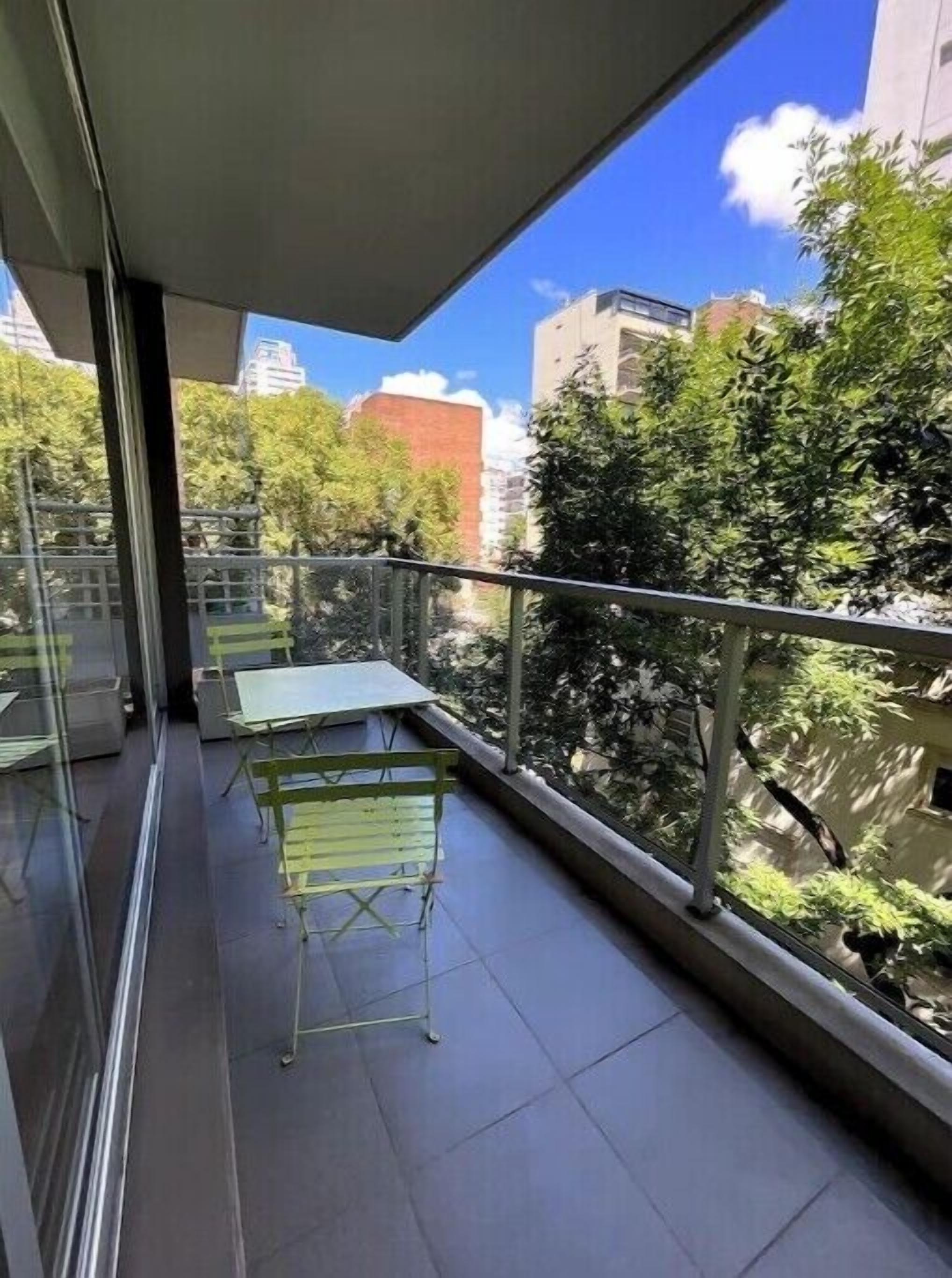 Un balcón con mesa y sillas, paredes de vidrio y vistas a árboles y edificios.