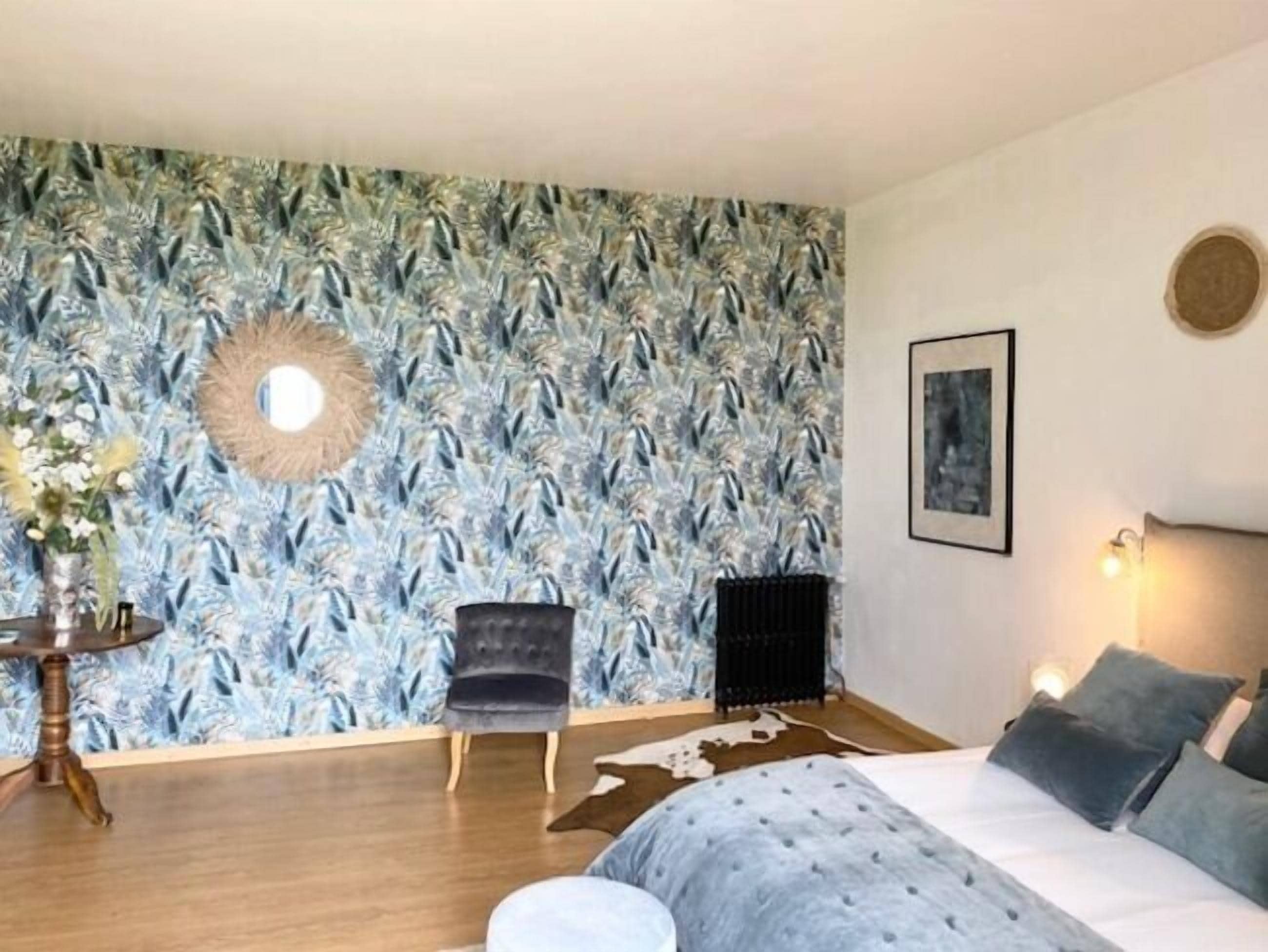 A bedroom with a patterned wallpaper, a round mirror, a framed picture, a bed with grey bedding, and a cowhide rug.