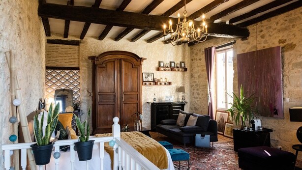 Una habitación rústica con vigas de madera a la vista, un candelabro, una chimenea, un sofá y una cama con cabecera blanca.