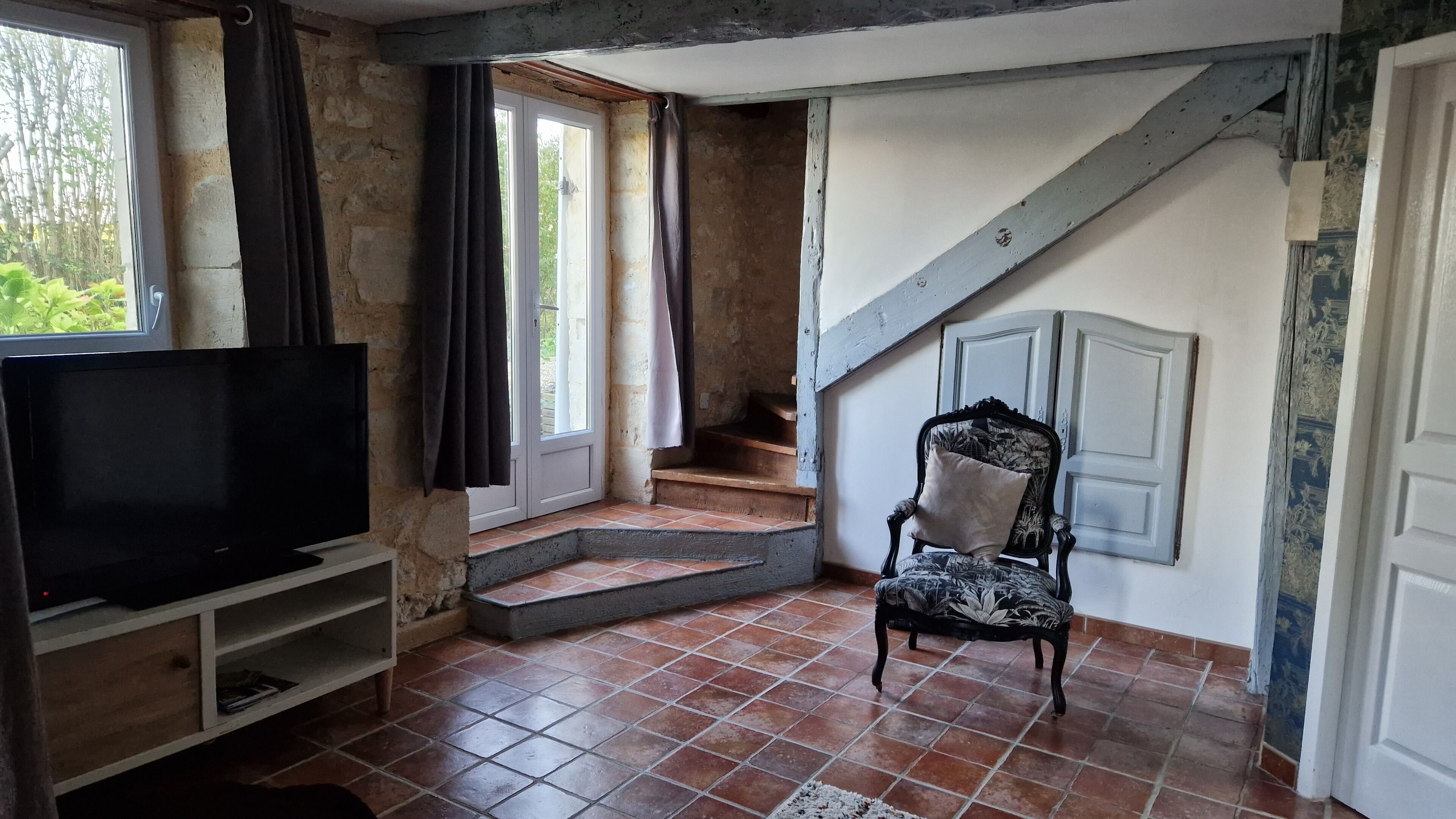 Ein Zimmer mit einem Fernseher auf einem Holzboden, ein Sessel mit einem gemusterten Kissen, eine Treppe und eine Tür mit floralem Dekor.