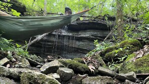 Eine Hängematte zwischen Bäumen in einer bewaldeten Gegend mit einem kleinen Wasserfall und Felsen.