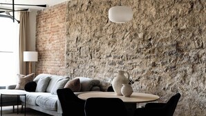 Un salon moderne avec un mur en briques, un canapé gris, des chaises noires, une table ronde et un luminaire suspendu blanc.