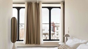 Une chambre d’hôtel avec une grande fenêtre, un lit avec du linge de maison blanc et une lampe sur une table de chevet.