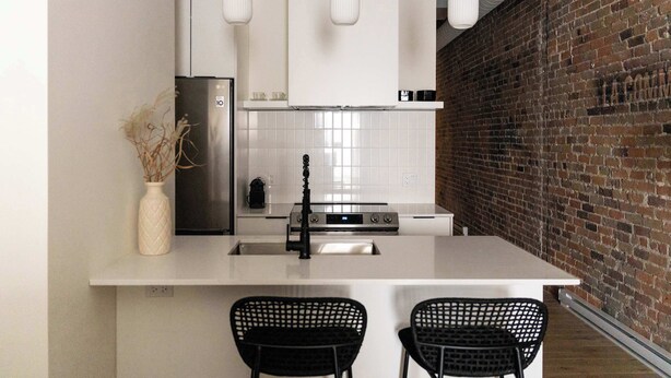 A modern kitchen with a white island, black chairs, a stainless steel refrigerator, and a brick wall. - Maison Place Jacques-Cartier • Luxury Loft • Old Montreal • Pet Friendly • Full Kitchen • Wi-Fi • AC (Montréal)