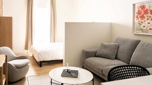 A modern living room with a grey sofa, a round coffee table, a grey armchair, and a bed with white bedding. A framed picture of flowers hangs on the wall. - Maison Place Jacques-Cartier • Luxury Loft • Old Montreal • Pet Friendly • Full Kitchen • Wi-Fi • AC (Montréal)