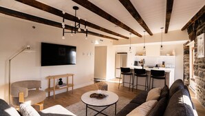Una sala de estar moderna con sofá, una mesa de centro y televisor. Se observa una zona de cocina con taburetes de bar y una pared de piedra.