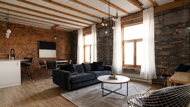 A modern living room with exposed wooden beams, a stone wall, a black sofa, a round coffee table, and a dining area with a white table and chairs. - Maison Place Royale • Luxury Waterfront One-Bedroom Apartment • Old Montreal • Water View St. Lawrence River • Full Kitchen • Stone Walls • Wi-Fi • AC (Montréal)