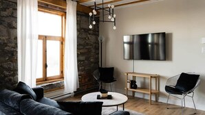 A modern living room with a stone wall, a wooden ceiling, a round coffee table, a black sofa, a TV, and a hanging light fixture. - Maison Place Royale • Luxury Waterfront One-Bedroom Apartment • Old Montreal • Water View St. Lawrence River • Full Kitchen • Stone Walls • Wi-Fi • AC (Montréal)