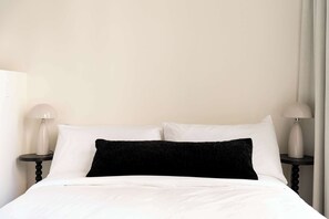 A neatly made bed with white linens and a black accent pillow, flanked by two bedside tables with lamps, in a simple room with plain walls. - Maison Place Jacques-Cartier • Luxury Loft • Old Montreal • Walk to Restaurants & Shops • Full Kitchen • Wi-Fi • AC (Montréal)