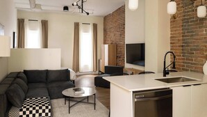A modern living room with a sofa, a coffee table, and a checkered ottoman. There is a kitchen area with a sink and a dishwasher. The room features a brick wall and a large window with curtains. - Maison Place Jacques-Cartier • Luxury Loft • Old Montreal • Central • Full Kitchen • Skylight • Wi-Fi • AC (Montréal)