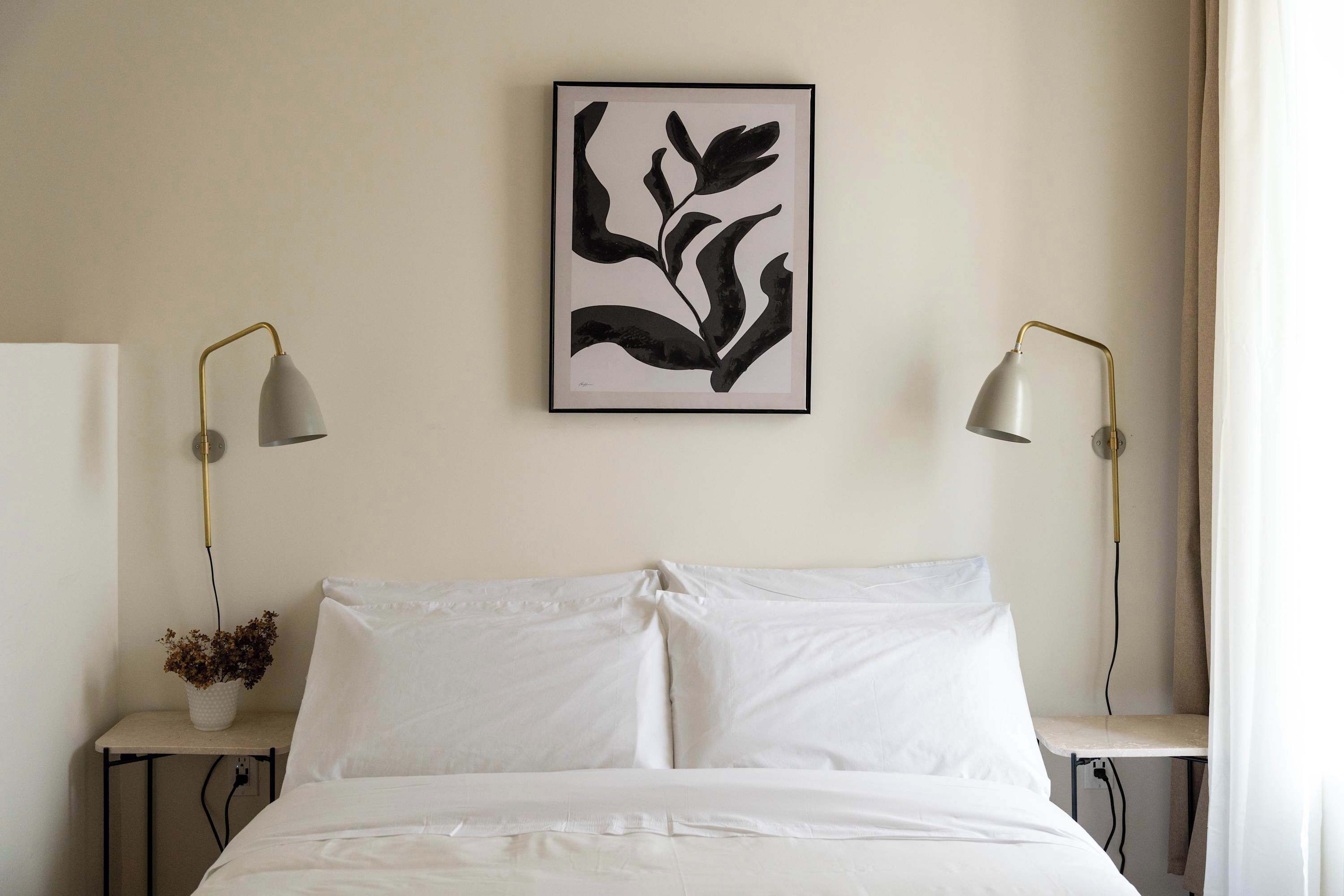 A neatly made bed with white linens, a framed abstract art piece above it, and two bedside tables with lamps on either side.
