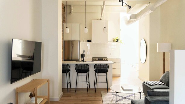 A modern living room with a flat-screen TV, a wooden side table, and a grey sofa. There is a kitchen area with a white countertop, three black bar stools, and hanging pendant lights. - Maison Place Jacques-Cartier • Luxury Loft • Old Montreal • Central • Full Kitchen • Skylight • Wi-Fi • AC (Montréal)