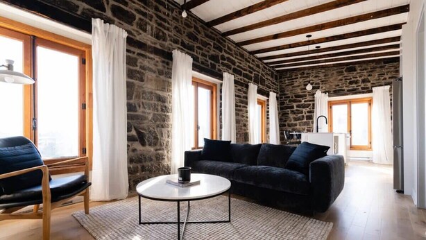 Un espacio habitable amplio con una pared de piedra, un sofá negro, una silla de madera y una mesa de centro redonda.