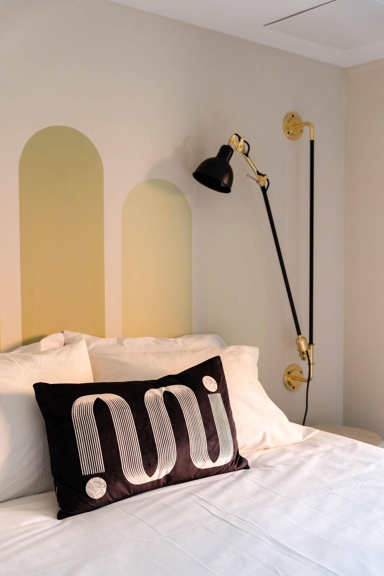 Un lit avec une literie blanche, un oreiller noir à rayures blanches et une lampe de bureau noire avec un pied doré.