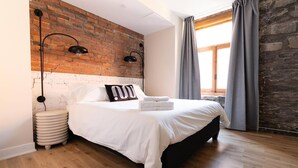 A bedroom with a brick wall, a bed with white bedding, two wall-mounted lamps, and a window with curtains. - Maison Place Royale • Luxury Two-Bedrooms Apartment • Old Montreal • Full Kitchen • Stone Walls • Wi-Fi • AC (Montréal)