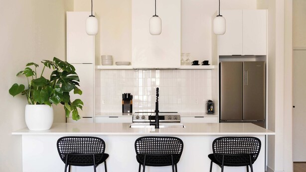 A modern kitchen with a white island, black bar stools, a potted plant, and stainless steel appliances.