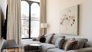 A modern living room with a grey sofa, a small round table, and a lamp. A large window with curtains is visible, and a framed artwork hangs on the wall.