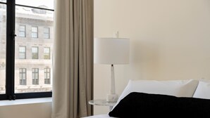 A hotel room with a bed, a nightstand with a lamp, a window with curtains, and a view of a building outside.