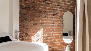 Ein Schlafzimmer mit Backsteinwand, einem Bett, einer Nachttischlampe, einem Spiegel und einem Fenster mit Vorhängen.