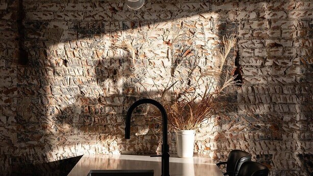 A kitchen with a stone wall, a stainless steel countertop, a black faucet, and a hanging light fixture.