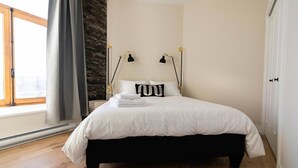 A bedroom with a bed, two bedside lamps, a window with curtains, and a stone wall.