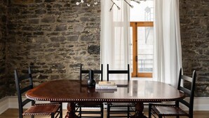 Un coin repas avec un mur en pierre, une table en bois, quatre chaises, un lustre et une fenêtre avec des rideaux.