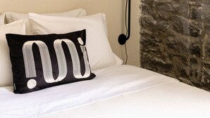 Un lit avec une literie blanche et un oreiller noir orné d’un motif blanc. Une lampe de sol noire est fixée au mur.