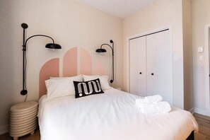 Una cama bien hecha con ropa blanca, dos lámparas negras fijadas a la pared, una puerta blanca y una pared con diseño geométrico.