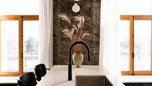 Una cocina con mostrador blanco, grifo negro, un florero con plantas secas y pared de piedra.