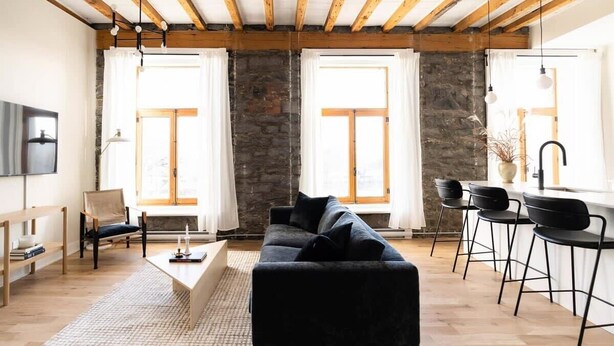 Una sala moderna con sofá negro, vigas de madera, pared de piedra y ventanas amplias.
