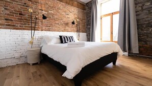 Un dormitorio con pared de ladrillo, una cama con ropa de cama blanca, dos lámparas de noche y una ventana con cortinas.