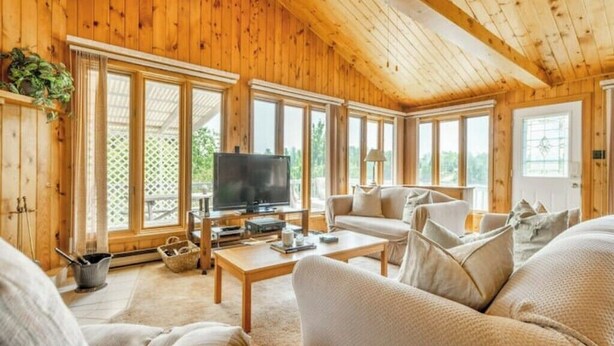 A cozy log cabin interior with a wooden ceiling, large windows, a television, and a comfortable seating area. - Jack's Lake Retreat - Perfect for multi-families! (Golden Valley)