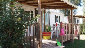 A wooden deck with a covered porch, a table with chairs, and a small garden area with a potted plant and a shovel. - ZEcamping 4* 500m plage parc aquatique dog friendly locatif TV clim (Hyères)