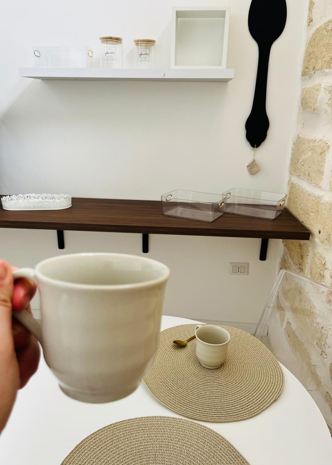 Una taza blanca sostenida por una mano, una pequeña taza sobre un tapete tejido y un recipiente transparente en un estante de madera.