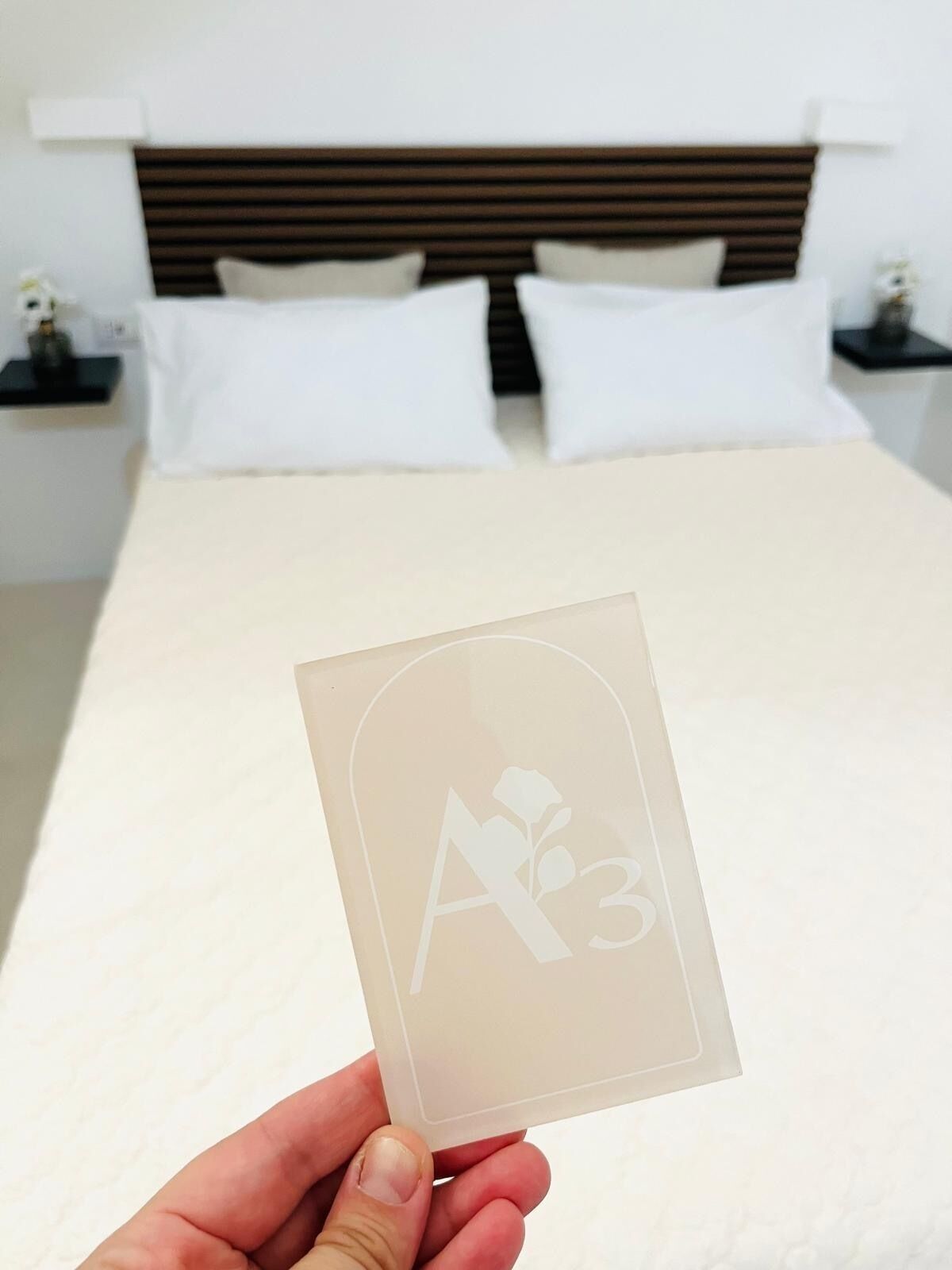 A hand holding a card with a rose and the number 3. In the background, there is a bed with white bedding and a wooden headboard.