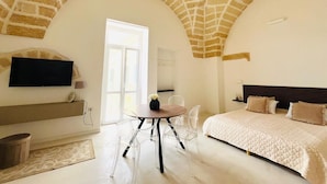 Ein Zimmer mit einer hohen Gewölbedecke, einem Bett mit gesteppter Bettdecke, einem an der Wand montierten Fernseher und einem Essbereich mit Tisch und Stühlen.