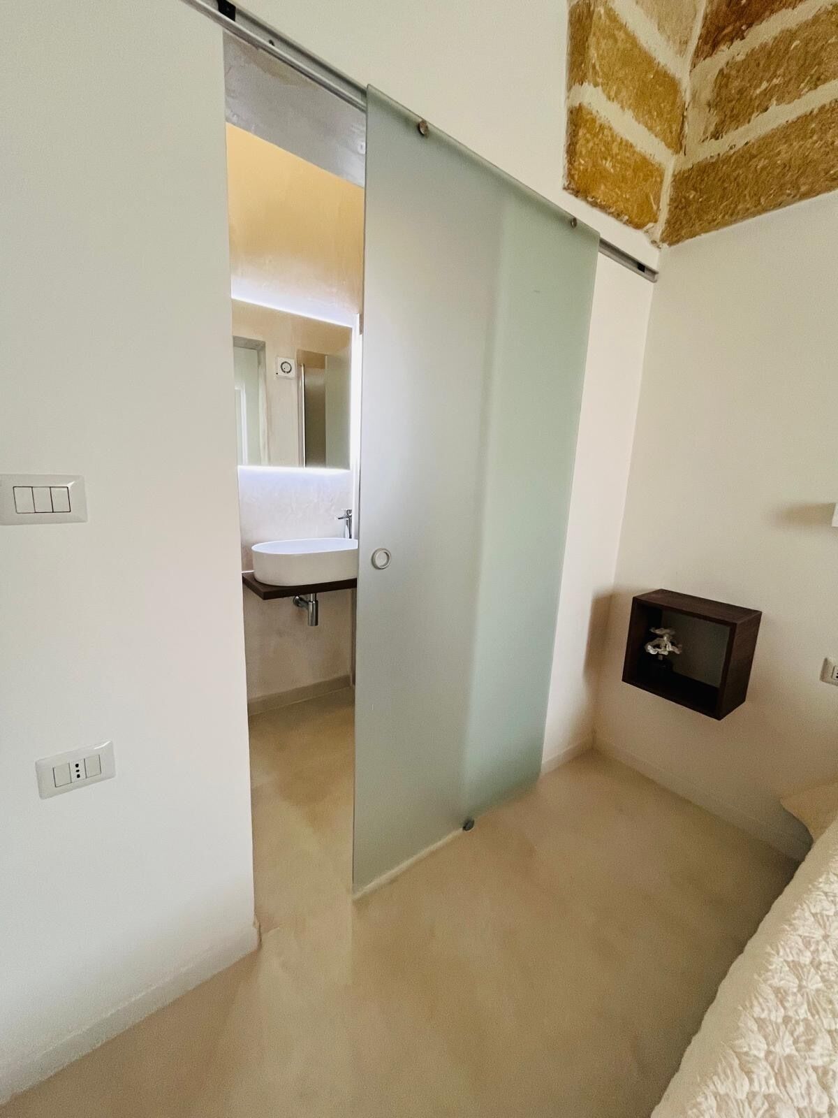 A bedroom with a sliding door leading to a bathroom. There is a small shelf with a decorative item on the wall.