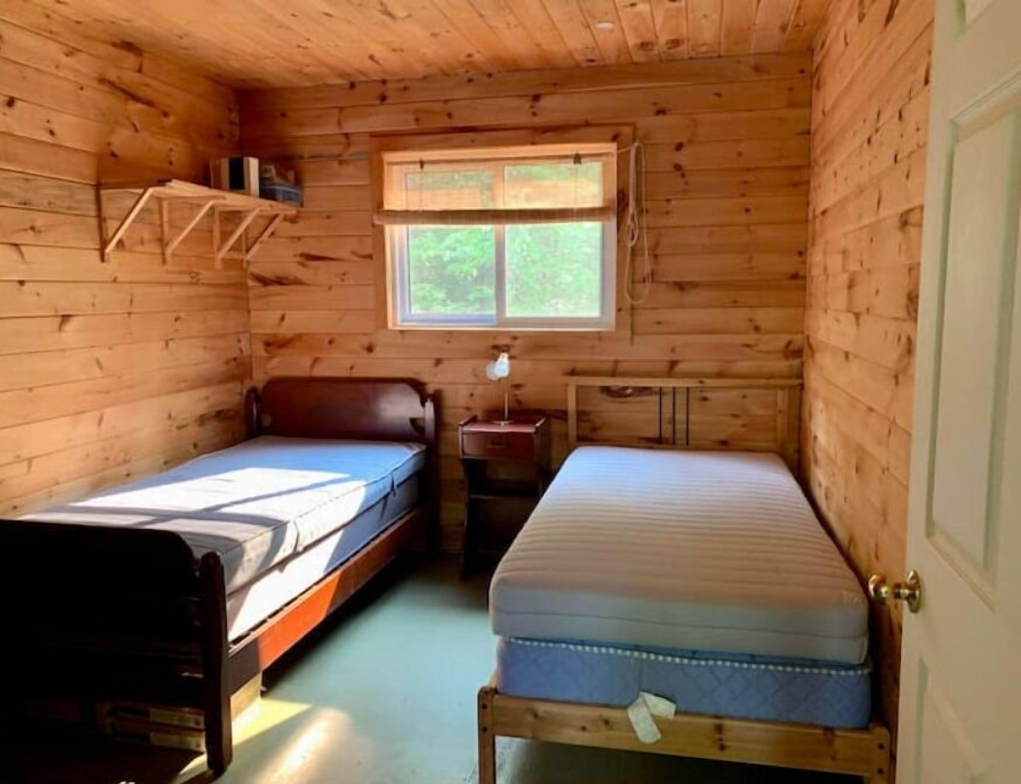 Una pequeña cabaña con dos camas individuales, una ventana con vistas a la vegetación y un interior de madera.