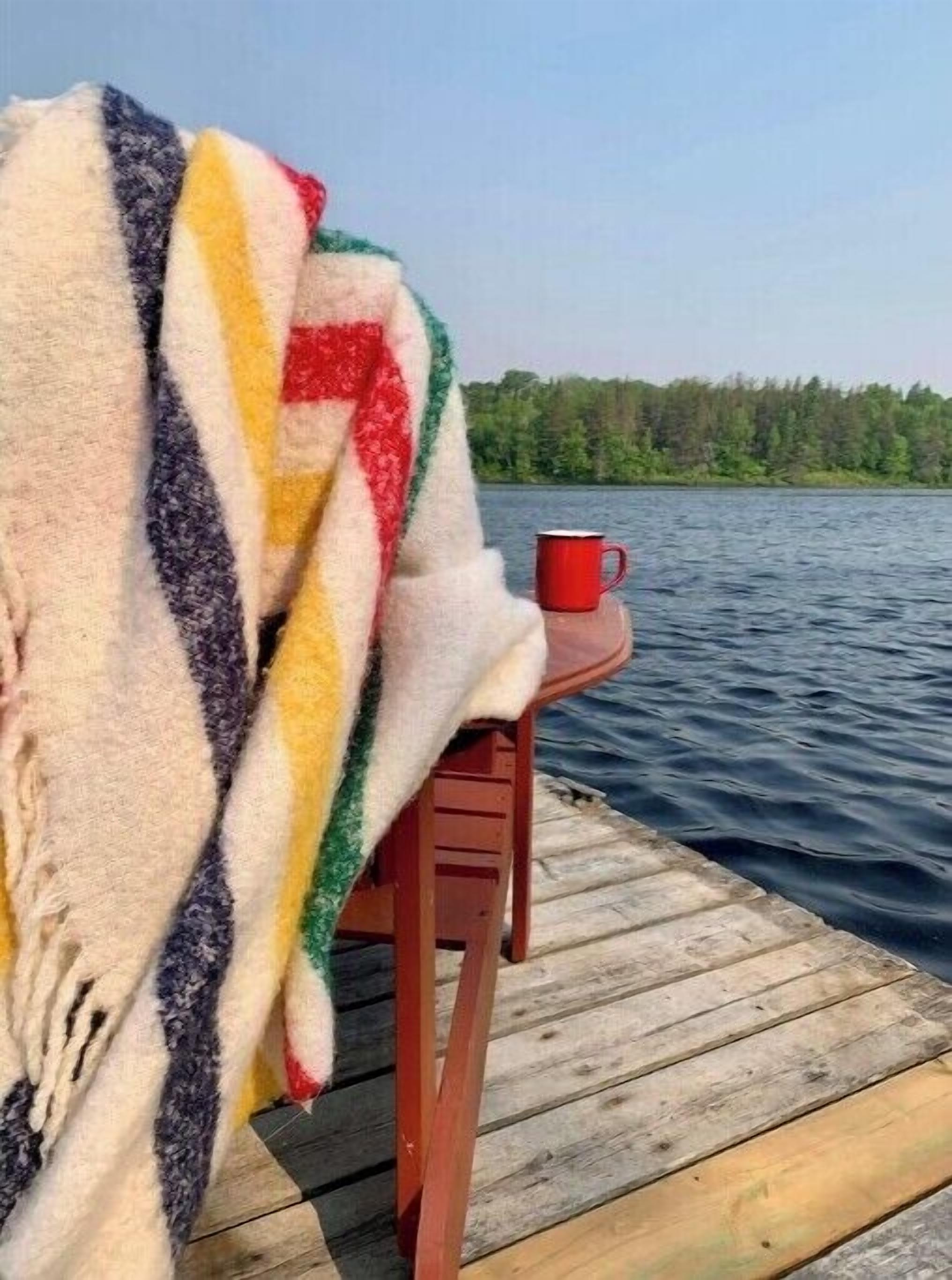 A wooden dock with a red chair and a red mug on it, overlooking a calm lake with trees in the background.