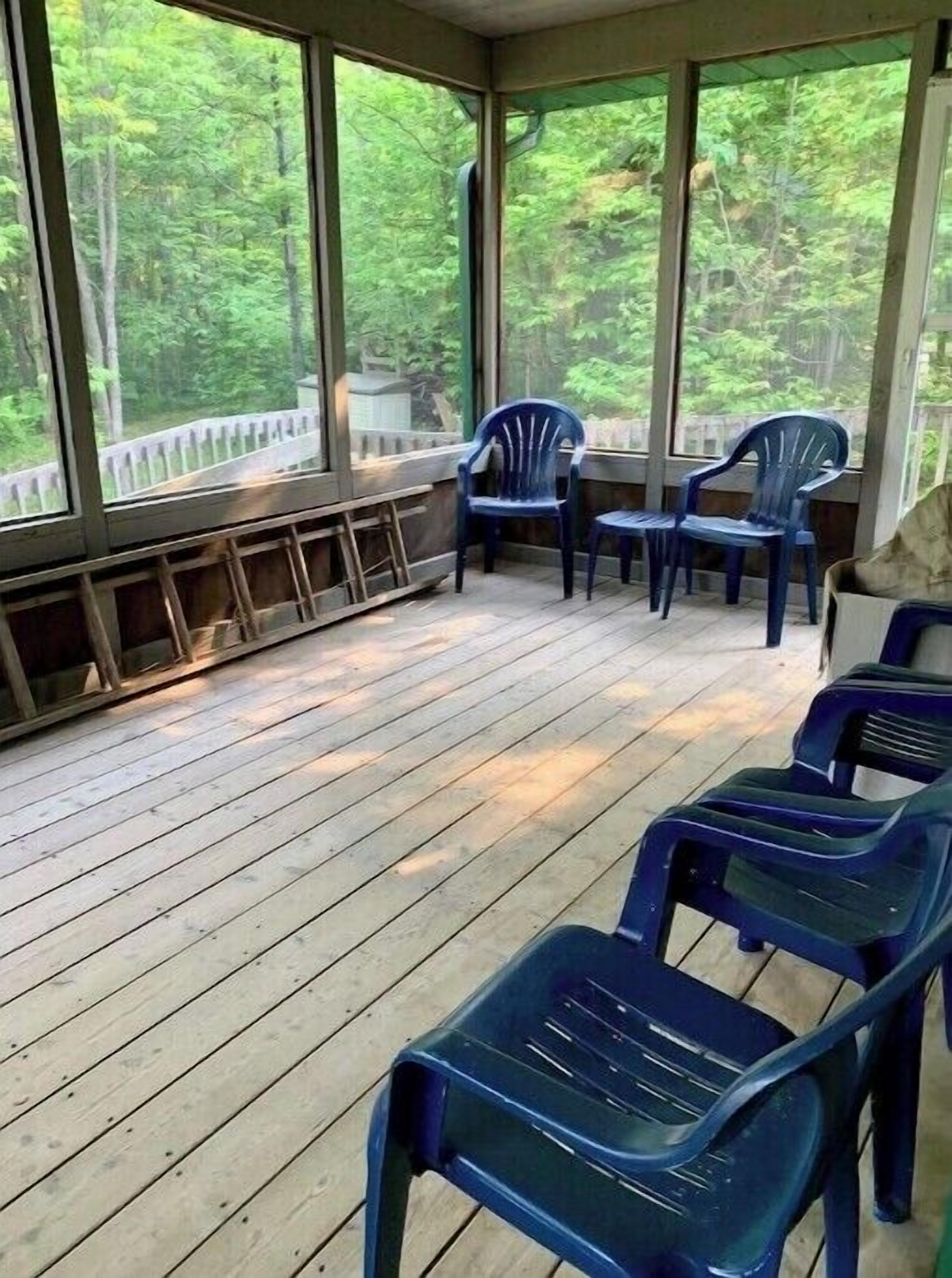 A covered outdoor area with wooden flooring and large windows offering a view of a forested area.