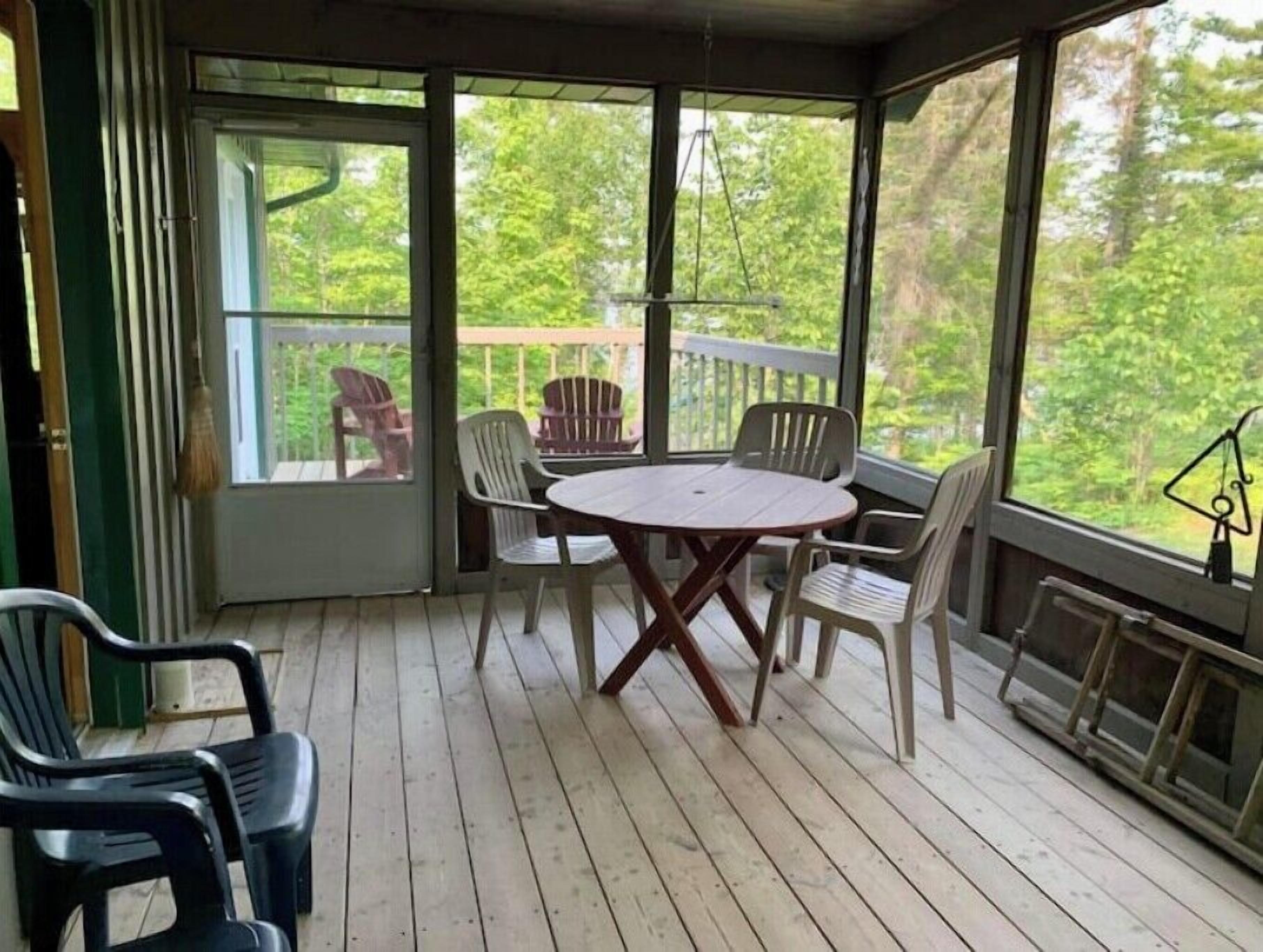 A covered porch with a round table and chairs, a view of trees through large windows, and a wooden deck.