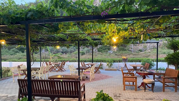 An outdoor patio with wooden tables and benches, surrounded by greenery and a pergola with hanging lights. - MERMAID PANSIYON (Marmaris)