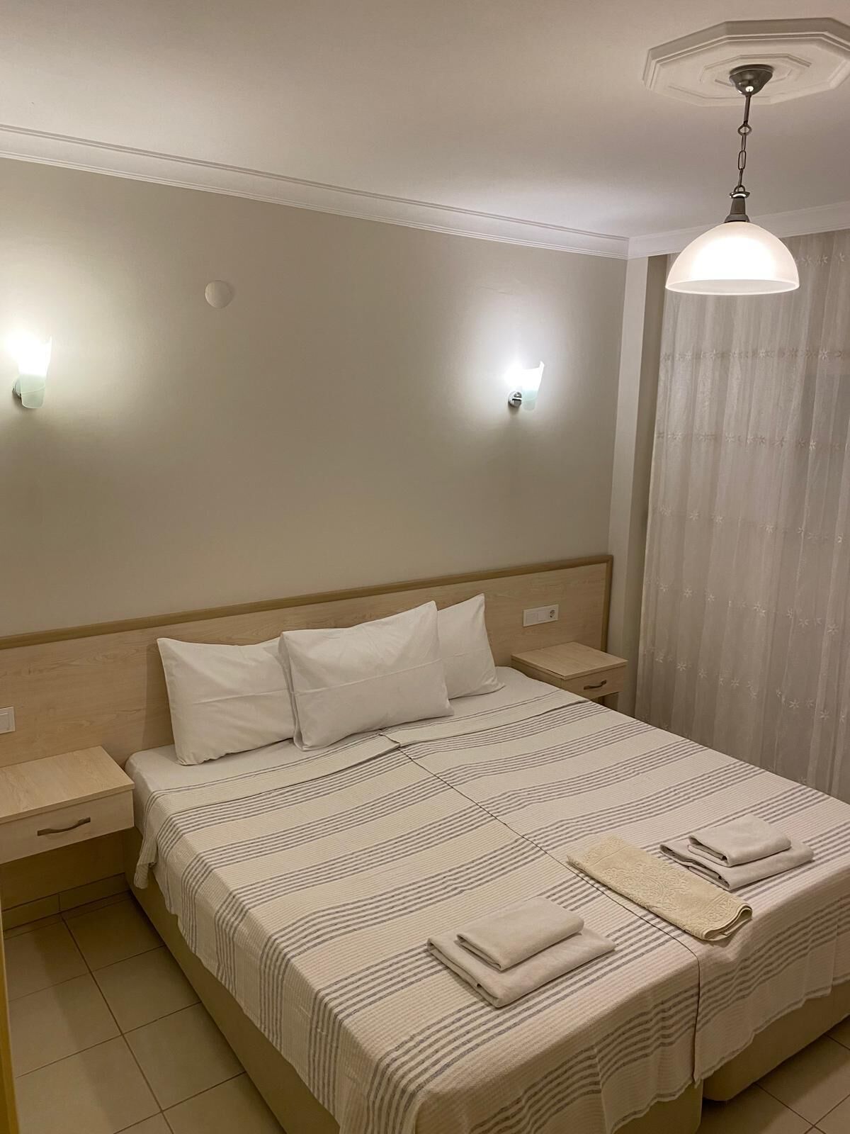 Una habitación de hotel con una cama grande, dos mesitas de noche, una lámpara de techo y un ventanal con cortinas.